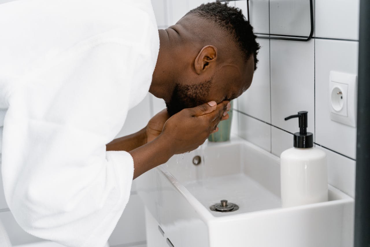 An adult man wearing a bathrobe washes his face at a sink, focusing on cleanliness.